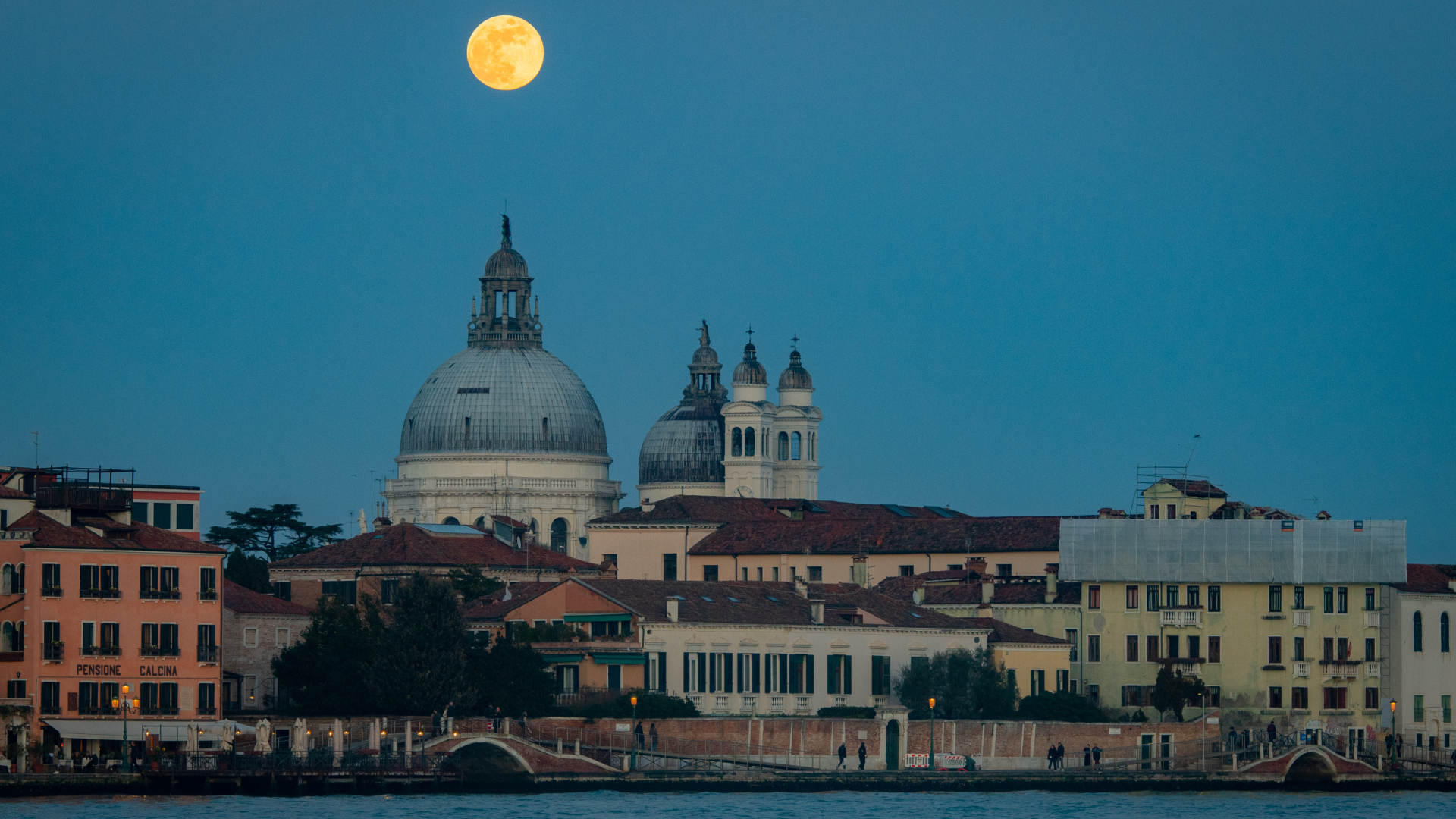 A yellow full moon is pictured above a city skyline dominated by the domed roof of a church in a darkening blue night sky. A body of water is visible in the foreground.