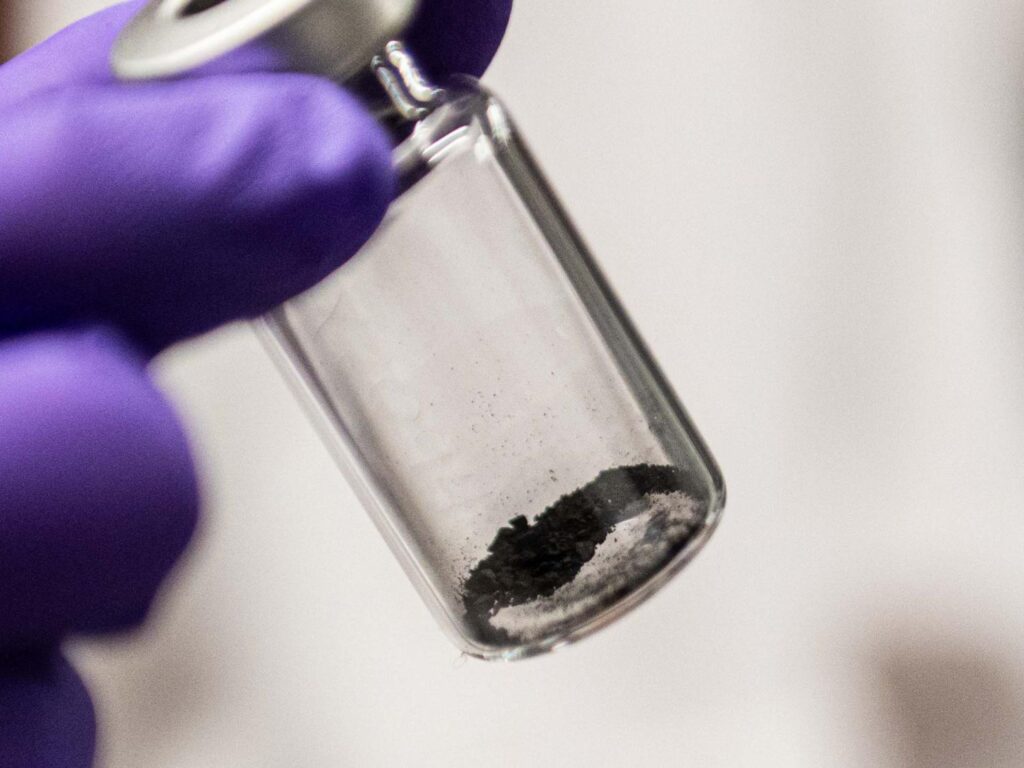 A purple gloved hand holding a clear tube with a little bit of dark gray dust within.