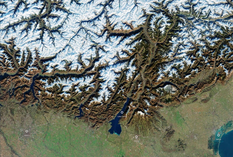 Satellite view of the snow-capped Alps mountains with lakes and green plains below; labeled locations include Lake Geneva and other geographic features.