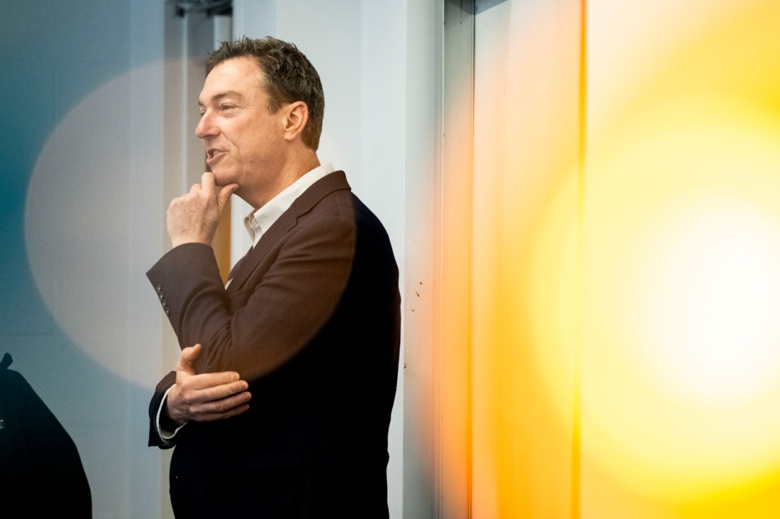 Professor Stefan Kautsch wearing a brown suit stands in thought with one hand propped against his chin and the other resting against his elbow.