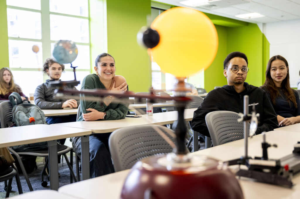 Students sitting in a classroom at tables watch a demonstration with an orrery (mechanical solar system model) in the foreground and a globe visible in the background. One woman sitting at the tables, wearing green, smiles broadly.
