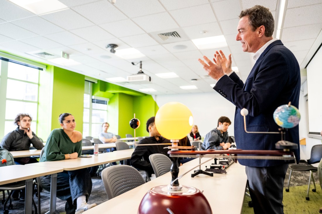 A professor gestures while lecturing to seated students in a bright classroom, with an orrery and globe on the table for demonstration purposes. 