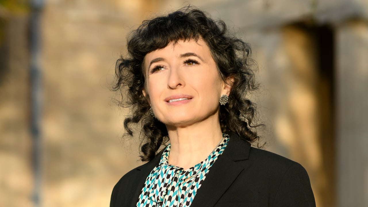 A woman with black curly hair wearing a checkered blouse and black blazer smiles off to the side of the camera. 