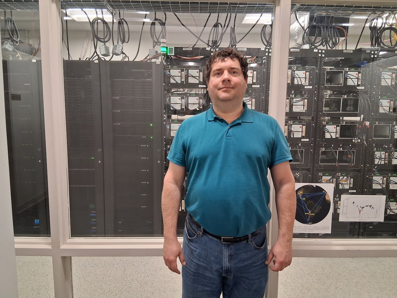 A man wearing a blue t shirt and jeans poses in front of stacks of computer software. 