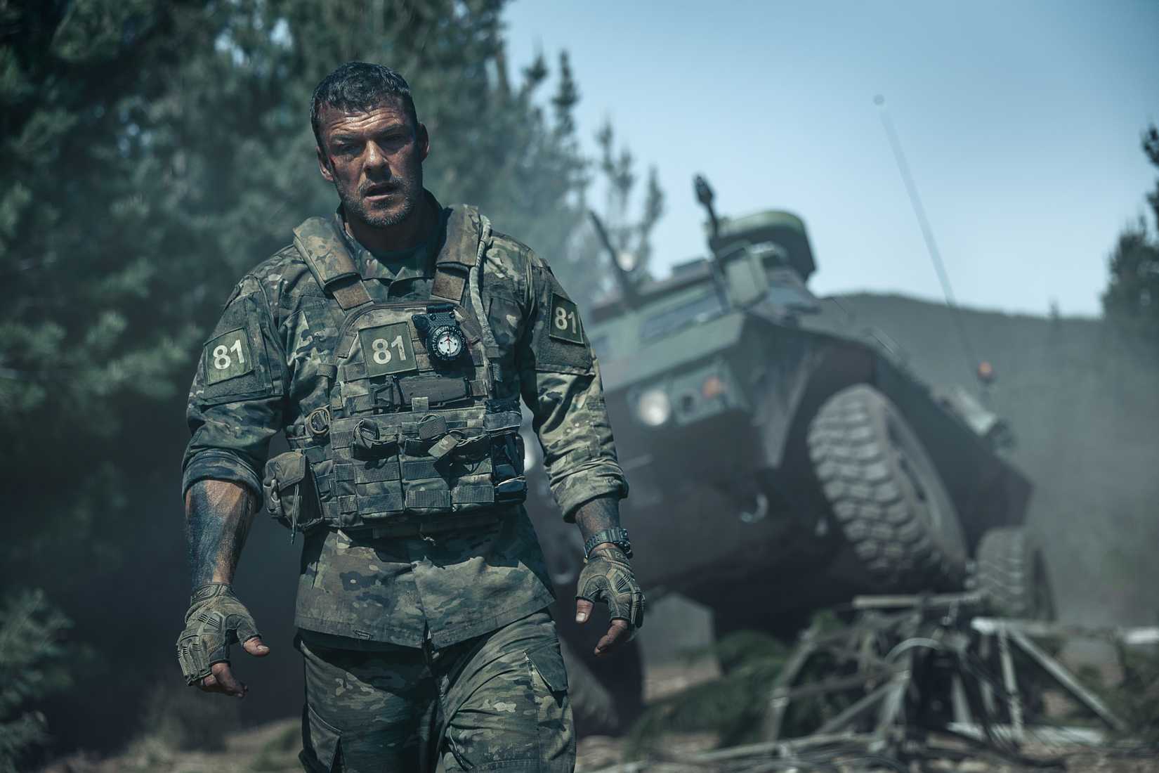 81 (Alan Ritchson), an Army sergeant in grimy fatigues, covered with mud and blood, stands in front of a wrecked army vehicle in War Machine