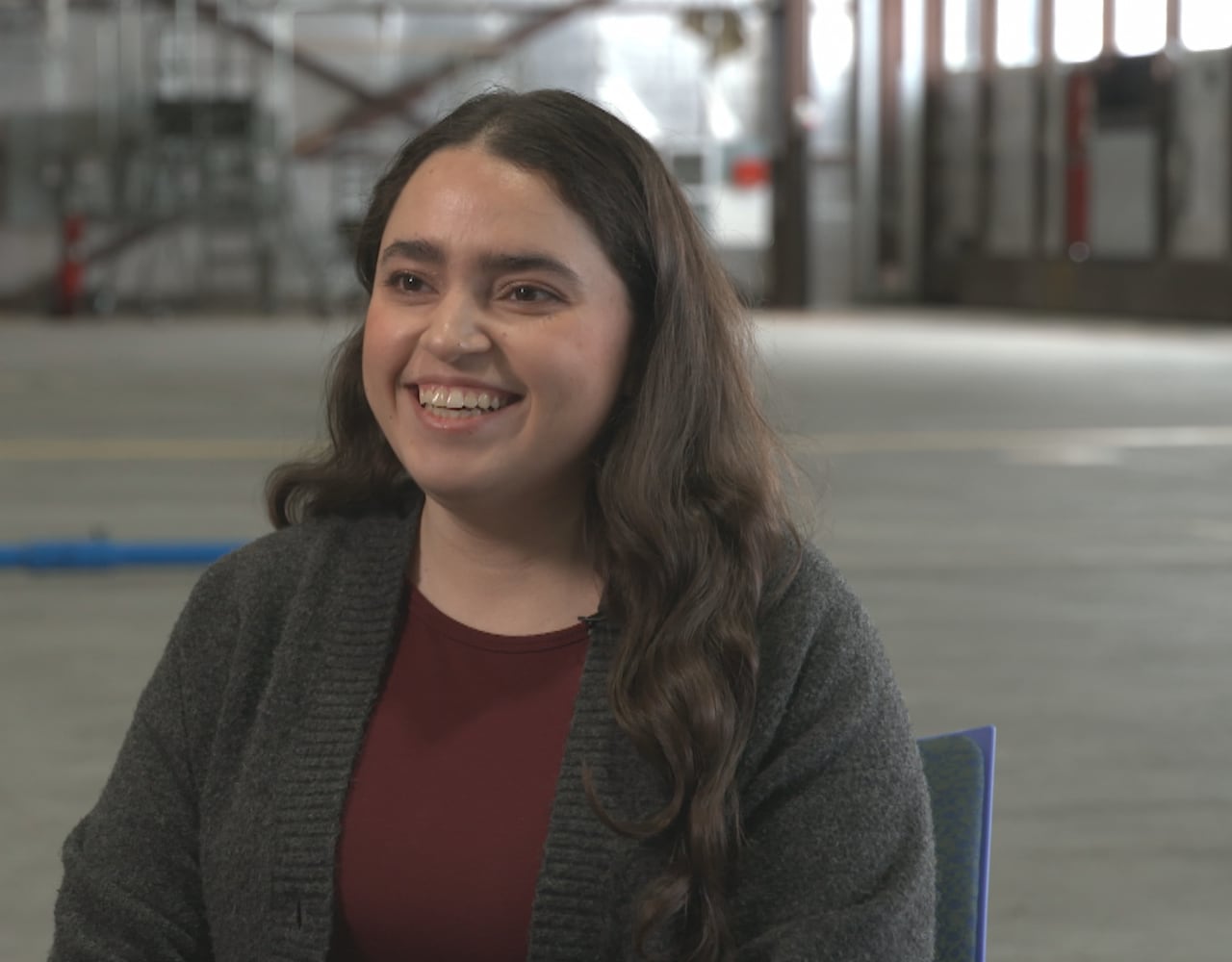A young woman with dark hair and a red shirt sits in a hangar. 
