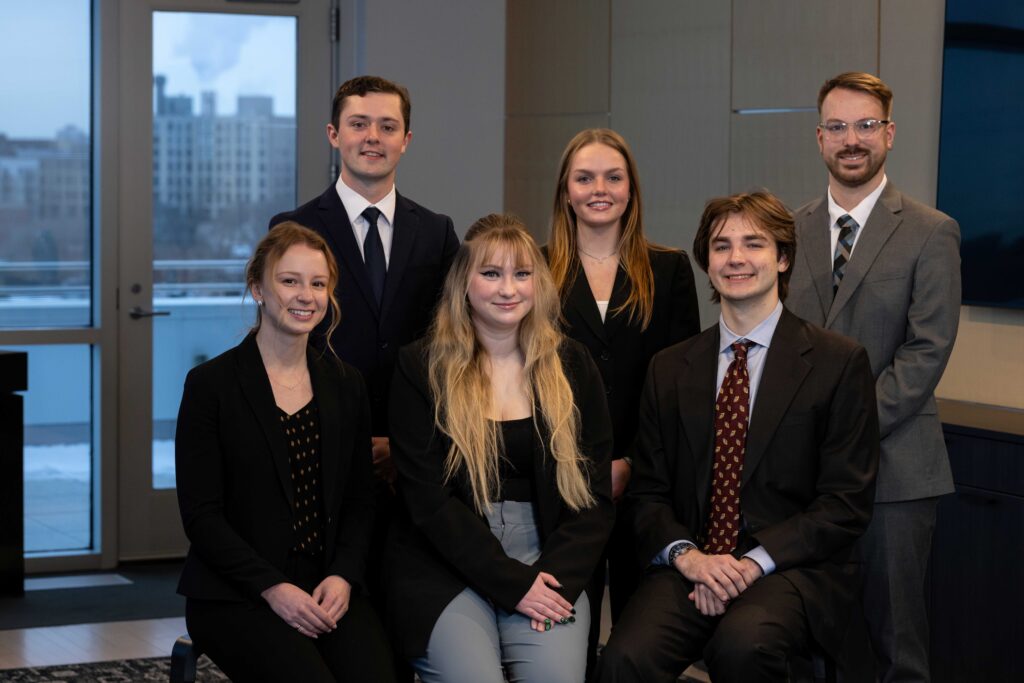 Six professionally dressed young adults pose together indoors, with three standing and three sitting in front. There are large windows showing a cityscape and cloudy sky in the background.