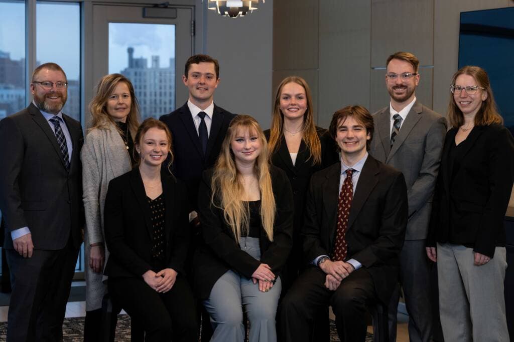 Nine professionally dressed people, six standing and three sitting, pose for a group photo in an office with large windows and city buildings visible in the background.