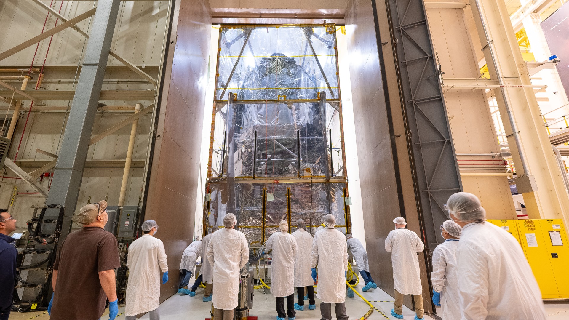 people in white lab coats look up at a large rectangular telescope sheathed in clear plastic in a large hangar