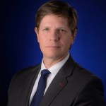 Portrait of Kevin Murphy, male, in front of a blue background wearing a black suit jacket and dark blue tie.