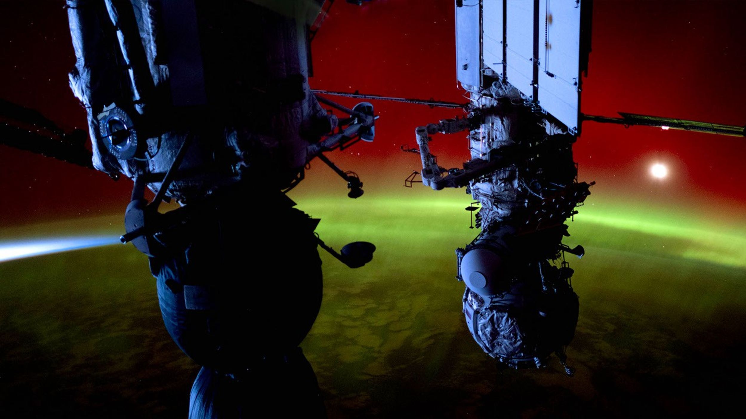 A striking photograph taken from the International Space Station in October 2024 showing spacecraft components silhouetted against a rare, intense red aurora with a band of green light below. The image captures parts of docked spacecraft or station modules in dark silhouette against the vibrant cosmic display, with a bright light visible in the upper right. The photograph dramatically illustrates astronaut Don Pettit's description of "flying in the aurora" rather than above it, as the station was immersed within this uncommon red auroral phenomenon at approximately 200+ miles altitude. The red glow, caused by oxygen atoms excited by an X1.8 solar flare that occurred on October 9, creates an otherworldly environment that, as Pettit described, made the ISS appear 'inserted into a neon sign.' This image documents the extraordinary geomagnetic storm that produced aurora visible from unusually low latitudes on Earth.