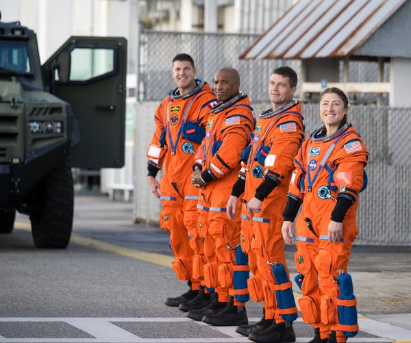 The four crew members of the Artemis II mission to the moon standing outside in orange spacesuits.
