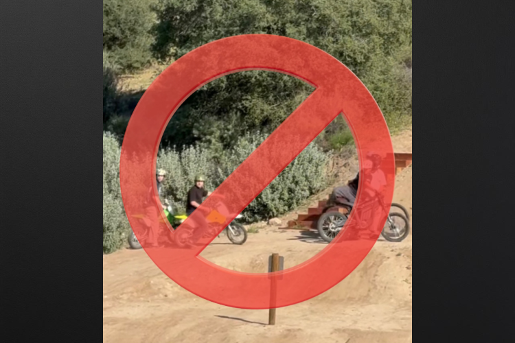 Two individuals on dirt bikes are positioned in a natural setting with greenery in the background. They appear to be observing or preparing for a ride, with a dirt path and a wooden ramp visible nearby. The image is overlaid with a red circle and diagonal line, indicating a prohibition of some sort, potentially suggesting that dirt biking is not allowed in this area.