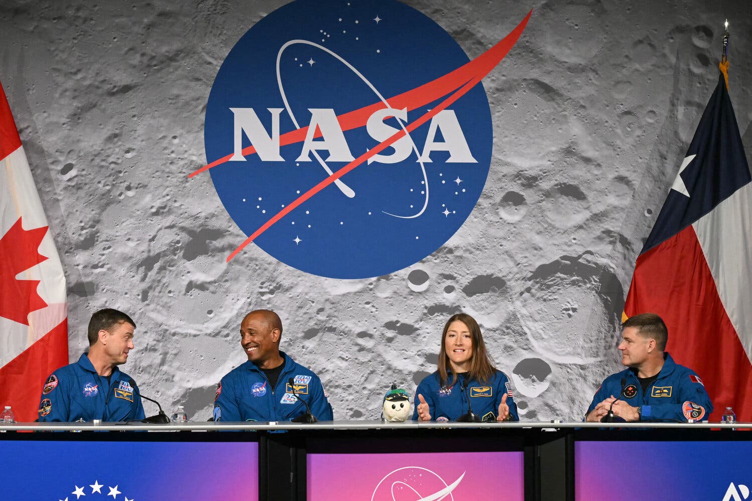 Four astronauts in blue jumpsuits on a stage with a large NASA logo superimposed over an image of the moon behind them.