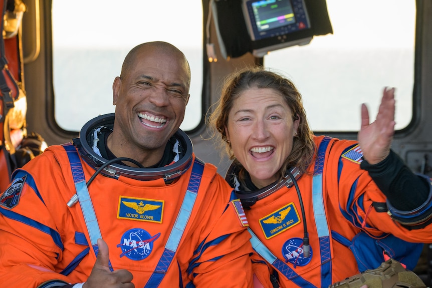 A man and a woman in orange NASA-branded space suits pose for the camera with a wave and a thumbs up.