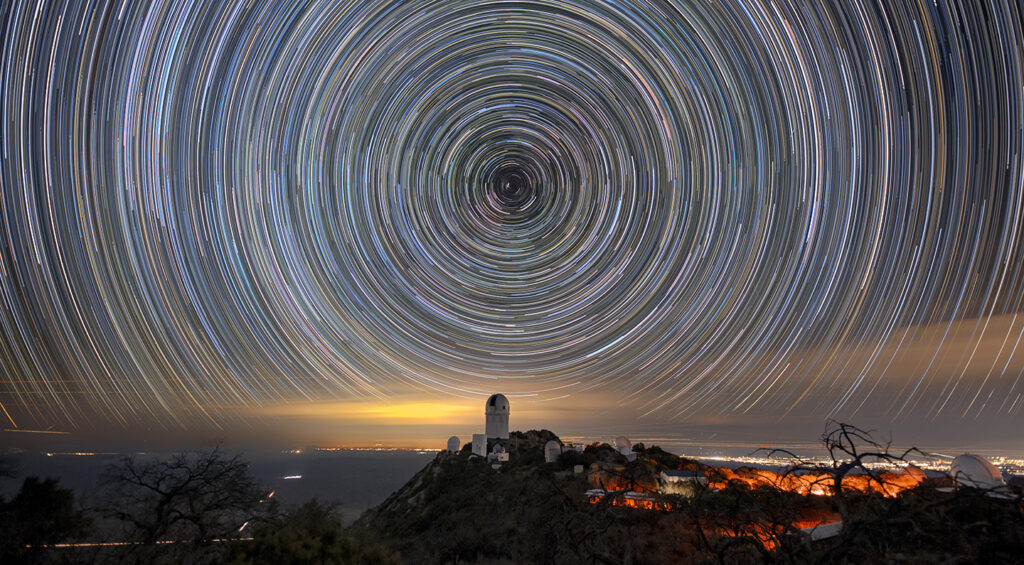 Circles of light on the night sky. A telescope dome atop a mountain is below the center of the circle.