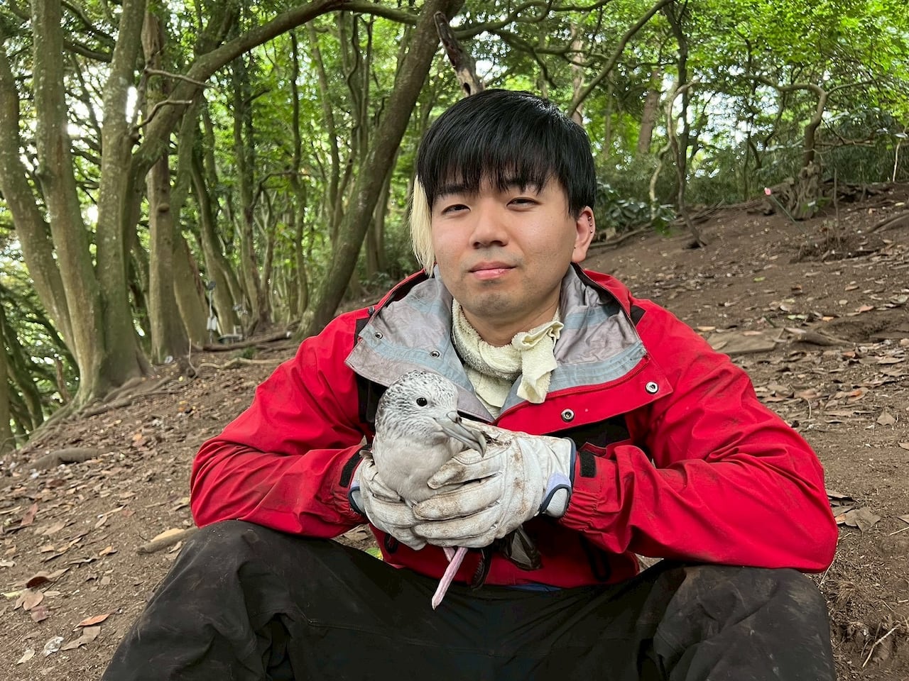 A man in a red jacket sits, while holding a seabird in his hands