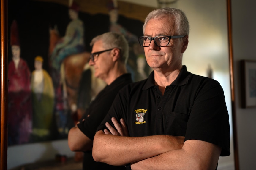 A man in a black shirt with black-rimmed glasses folds his arms standing in front of a mirror