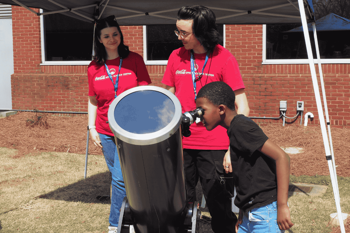  Gemini said A young boy looks through the eyepiece of a large solar telescope at an outdoor event. Standing behind the telescope are two Coca-Cola Space Science Center staff members wearing red t-shirts and blue lanyards, who are watching him with smiles. They are positioned under a white canopy tent in front of a brick building. The telescope has a large, blue-tinted solar filter on the front end.