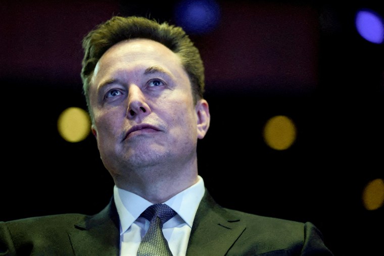 A close view of Elon Musk, seated in a darkened auditorium with colored lights behind him. 