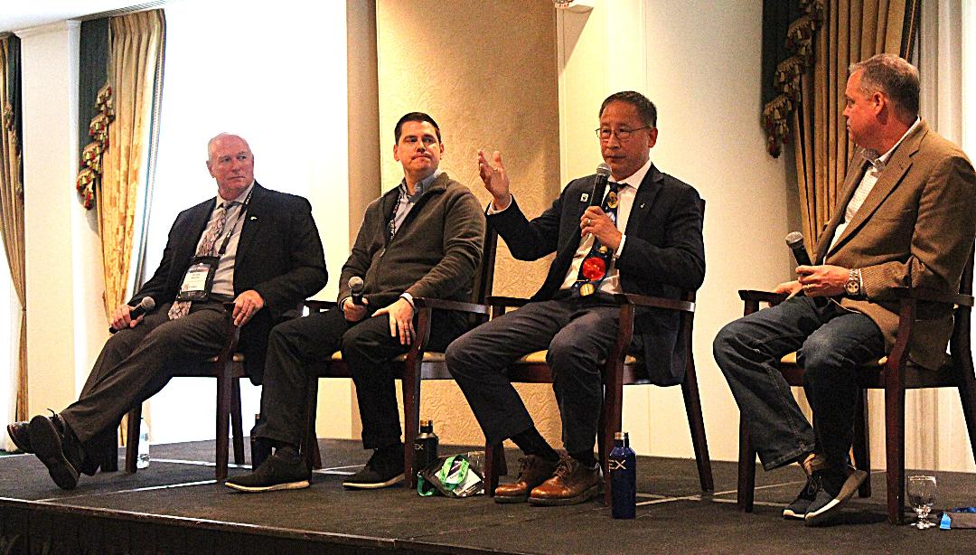 four people in suits talk into microphones on a stage