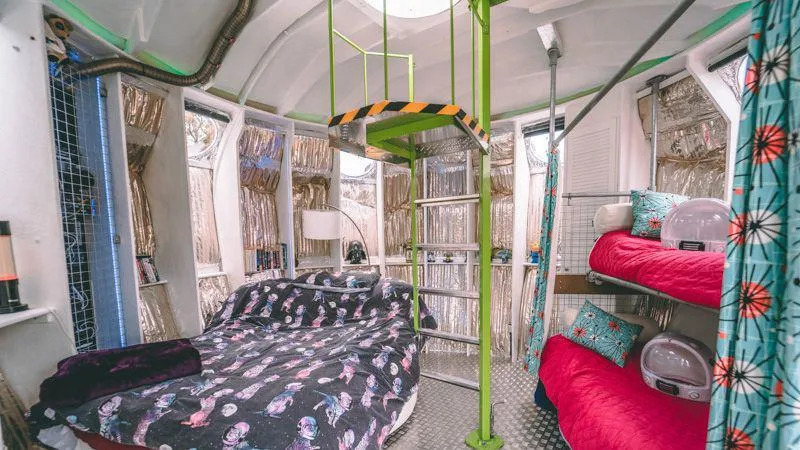 A look inside the pod's bedroom area. To the left is a double-bed with intergalactic themed bedding, above it is a lime-green stairway to an open top. To the right is a set of bunk beds with astronaut helmets perched on top. The floor is metal, there are metal tubes on the ceiling and aluminium shutters.
