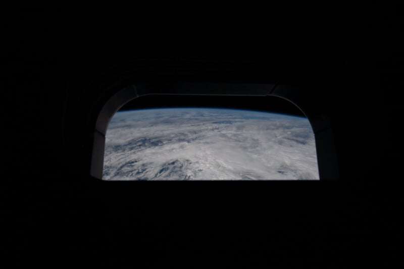 View of Earth's cloudy surface and blue horizon through a dark, rounded window, possibly from an airplane or spacecraft, with the surrounding interior mostly in shadow.