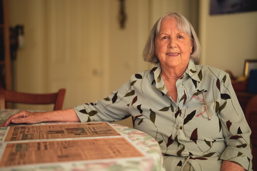 A woman aged in her 60s with grey bob hairstyle seated at a table, a newspaper in front of her