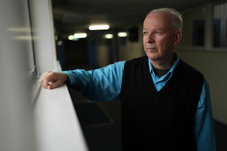 A man wearing a collared blue shirt and black vest leans on window sill looking outside
