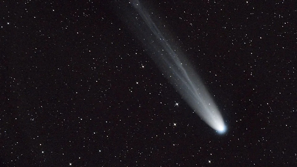 A streak of white across a dark starry night sky shows a comet moving toward the bottom right of the image. 
