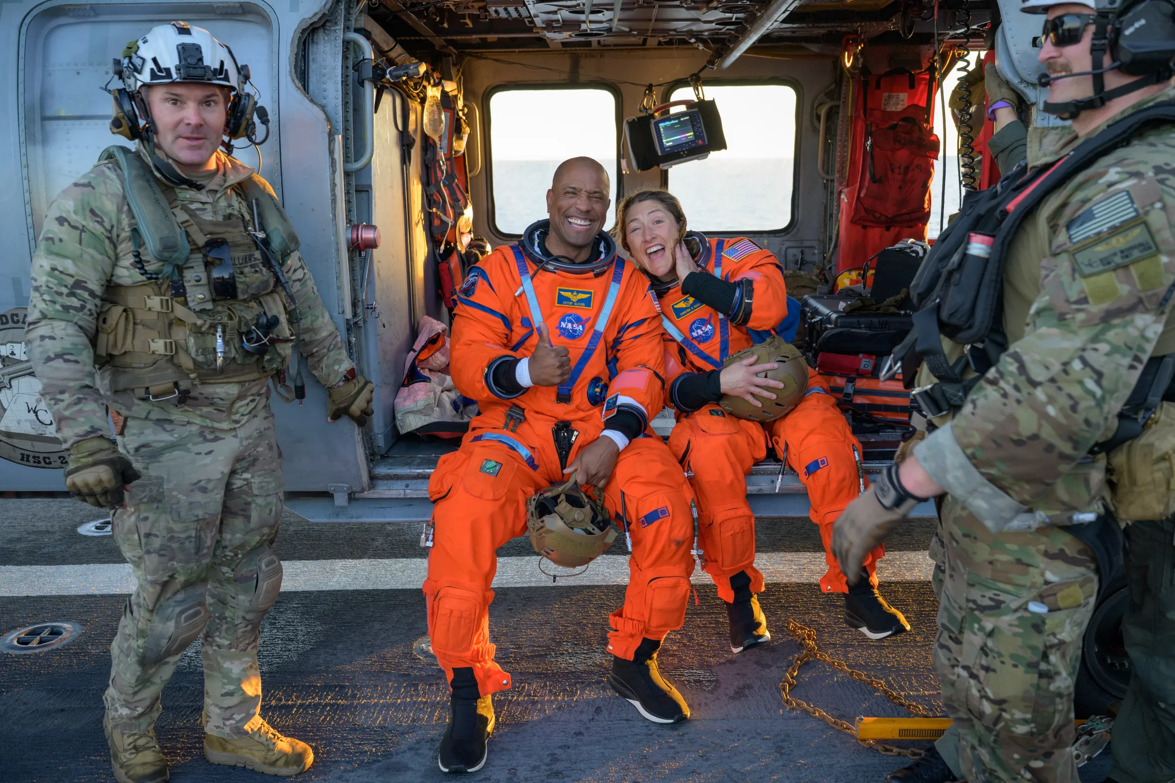 NASA astronauts Victor Glover and Christina Koch, wearing orange flight suits, pose for a photo inside a Navy MH-60 Seahawk helicopter.