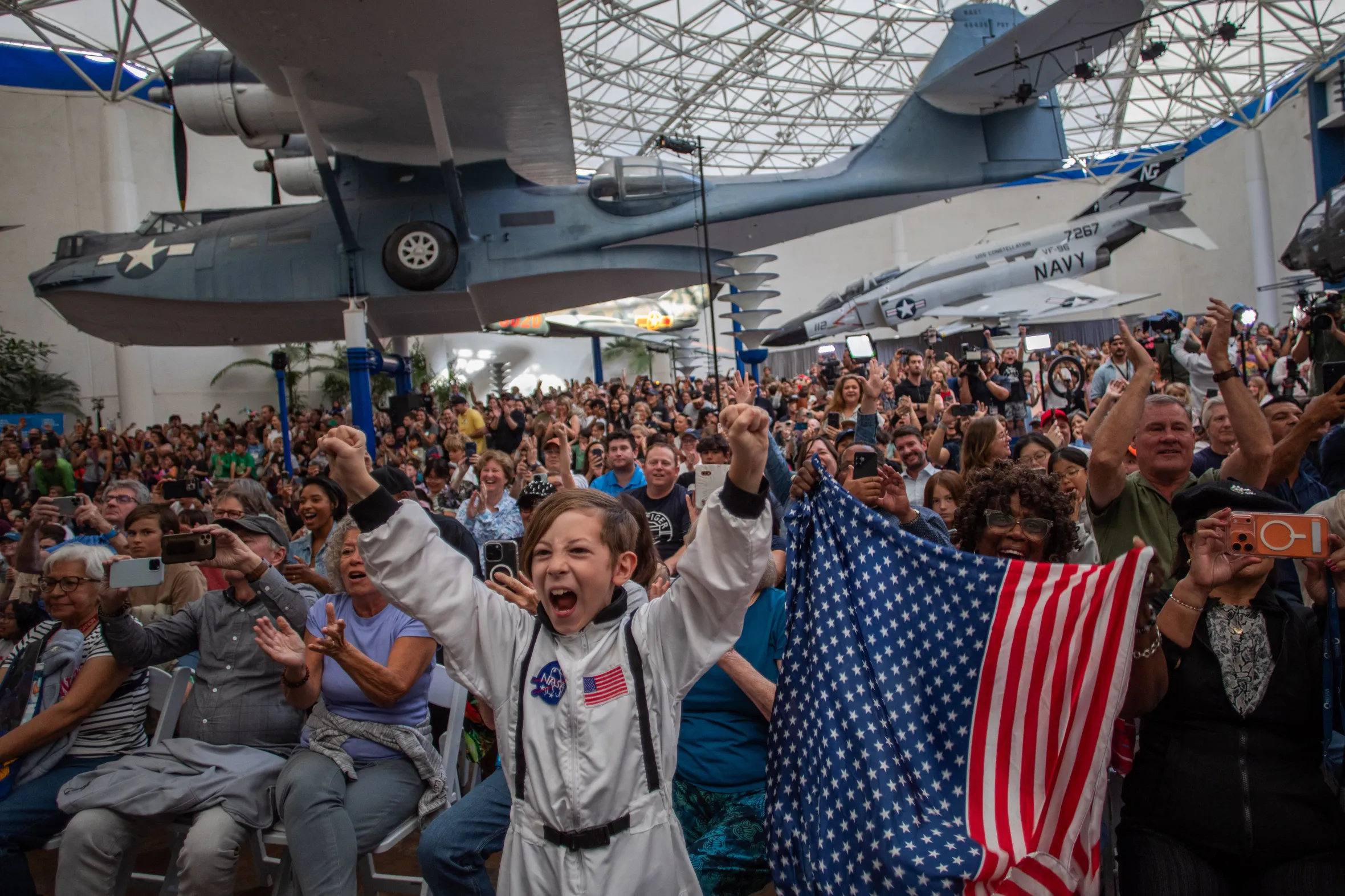 A boy in an astronaut costume cheers alongside a crowd waving an American flag at a live broadcast.