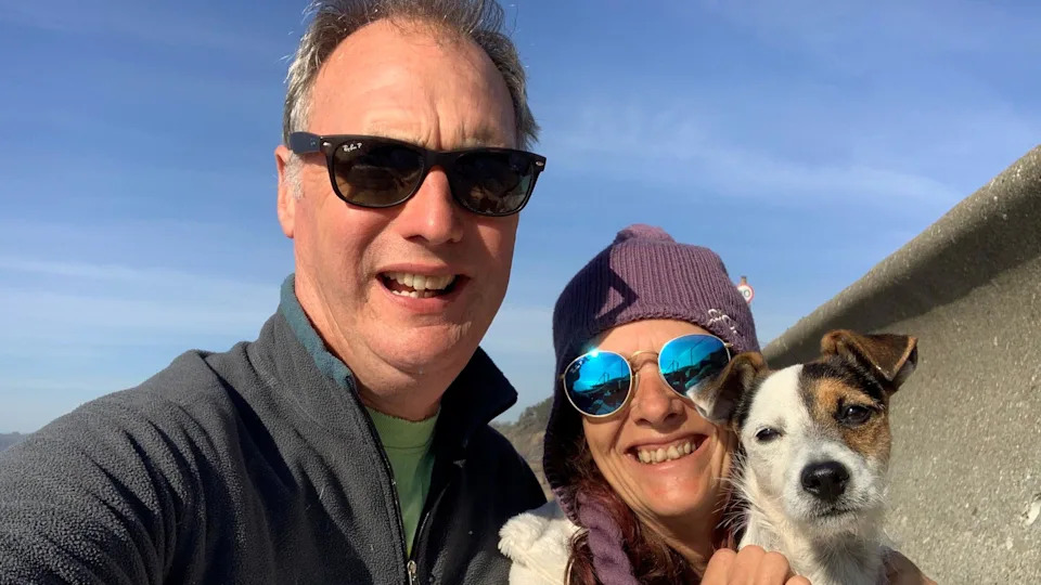 A grey-haired man with black sunglasses, wearing a blue fleece and holding his arm out to take a photo in selfie-mode. To his right, a brunette woman, wearing a purple hat and blue sunglasses. She holds her Jack-Russell dog in both hands. 