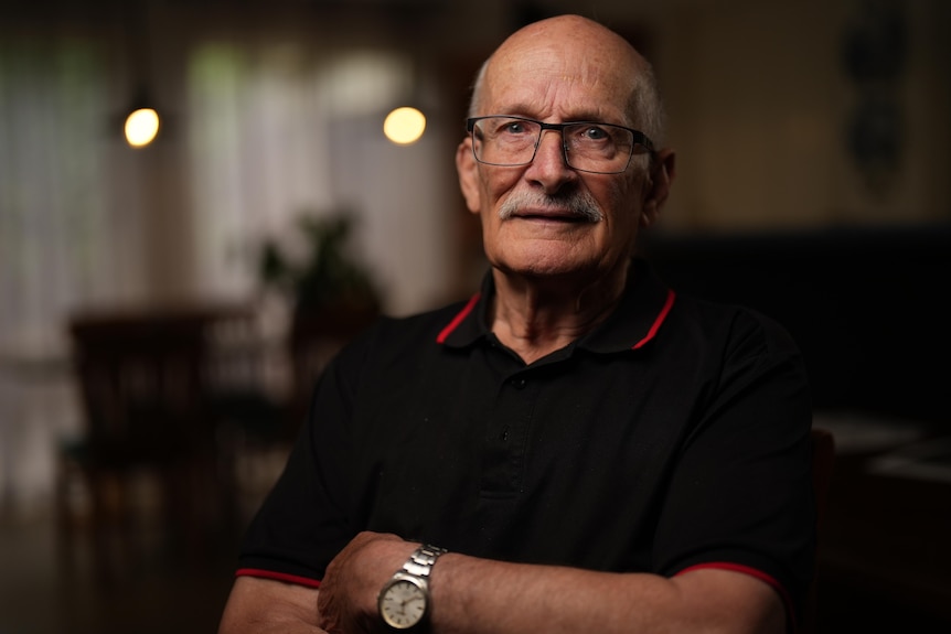A man in his 60s bald with a moustache and wearing glasses seated with arms crossed