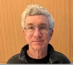 Headshot of a man with tousled grey hair and glasses, wearing a dark jacket.