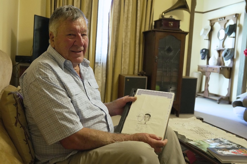 Elderly man seated on a loungeroom couch holds a photo of himself in the 1960s