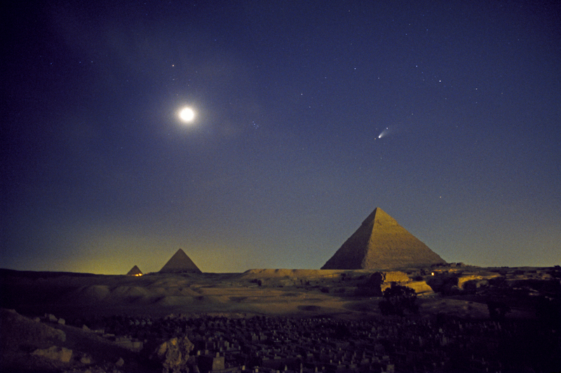 Bright round full moon and a comet in deep blue sky over a desert landscape with 3 enormous pyramids.