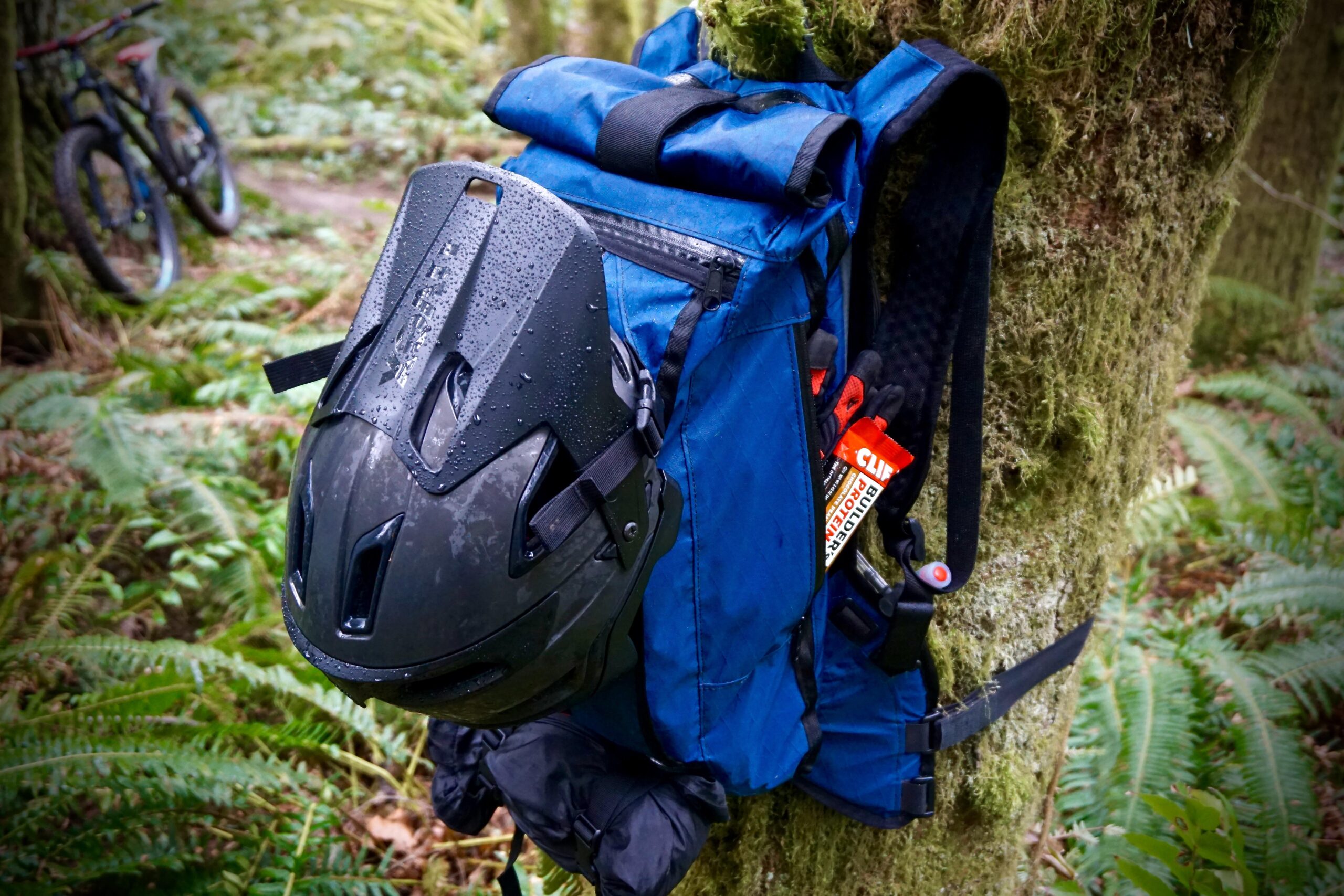 Mission Workshop The Hauser: A black bicycle helmet hangs from a blue backpack, which is secured to a tree in a lush, green forest setting. The backpack features a roll-top closure and has raindrops visible on its surface. Nearby, a mountain bike is partially visible, resting on the forest path. Various items, including energy bars, are accessible from the side pockets of the backpack.