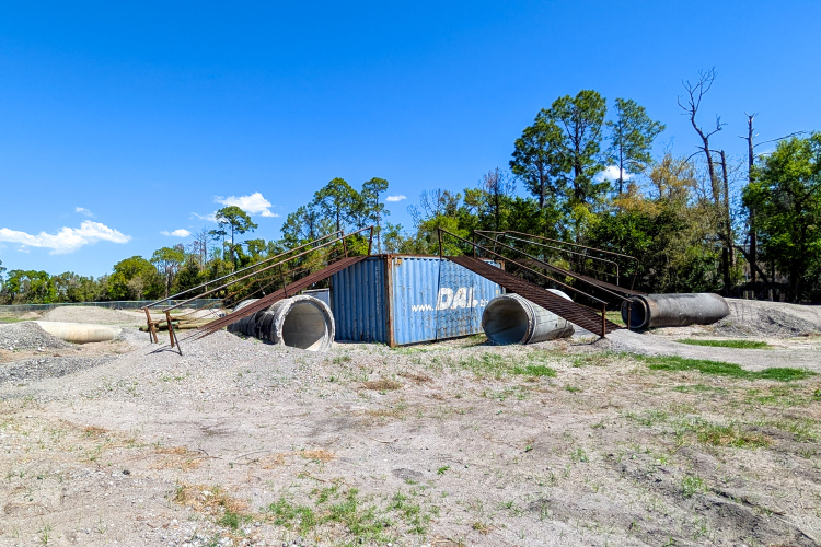 A rusted metal ramp leads up to a blue shipping container placed between two large concrete pipes, surrounded by a sandy, barren landscape dotted with sparse vegetation and patches of grass under a clear blue sky.