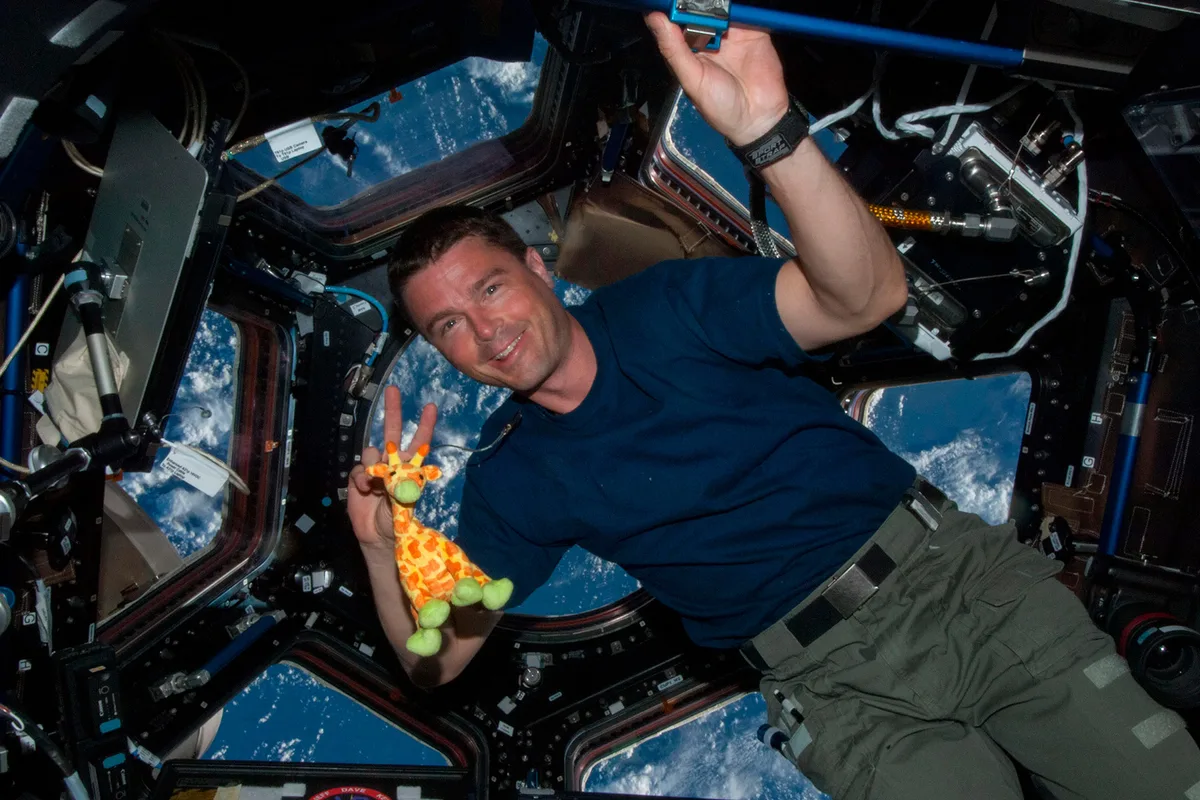 Reid Wiseman in the cupola with a toy giraffe with Earth in the background