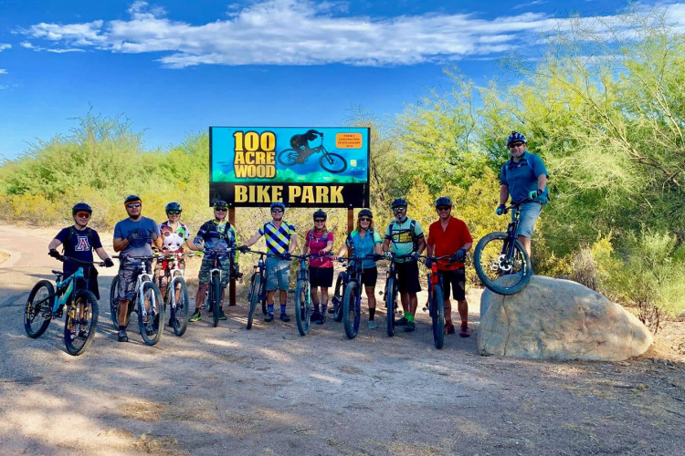 A group of ten people dressed in biking gear stands in front of a sign that reads "100 Acre Wood Bike Park." They are positioned on a dirt path surrounded by greenery, with some holding their mountain bikes. The sky is clear and blue with a few clouds, and one person is playfully posed on a rock with their bike.
