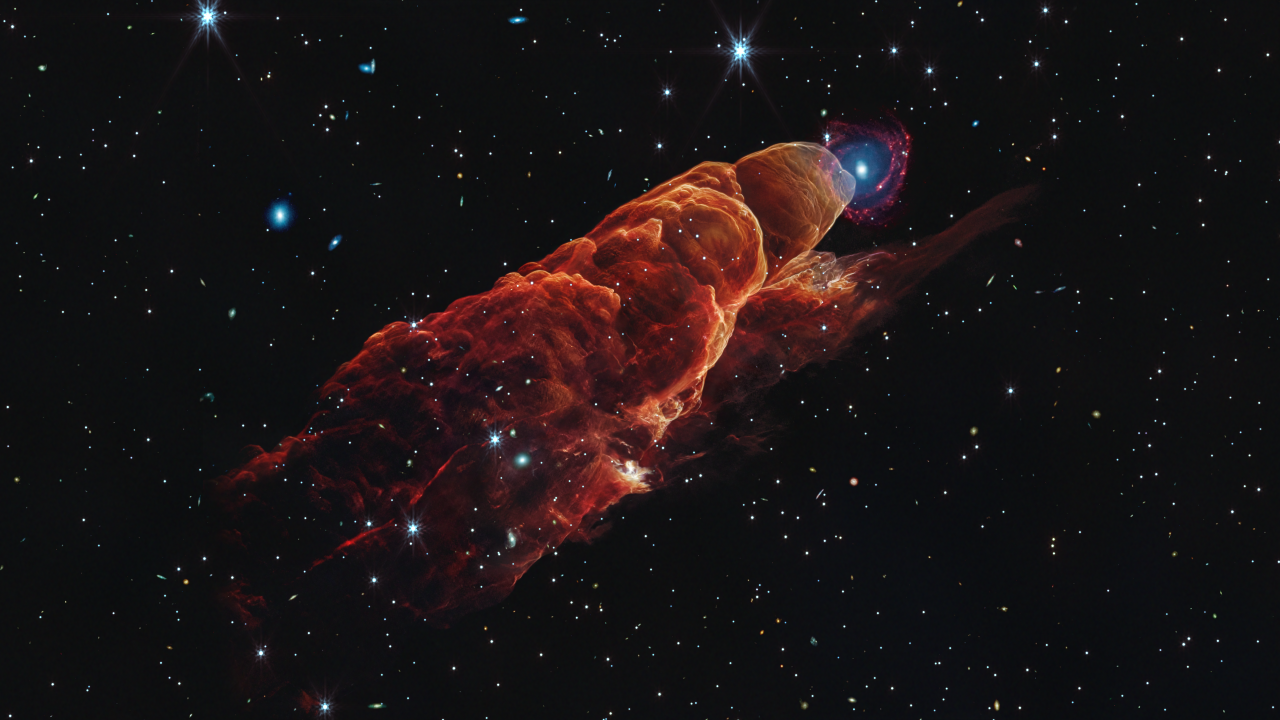 Angled from the upper left corner to the lower right corner is a cone-shaped orange-red cloud known as Herbig-Haro 49/50. This feature takes up about three-fourths of the length of this angle. The upper left end of this feature has a translucent, rounded end. The conical feature widens slightly from the rounded end at the upper right down to the lower right. Along the cone there are additional rounded edges, like edges of a wave, and intricate foamy-like details, as well as a clearer view of the black background of space. In the upper left, overlapping with the rounded end of Herbig-Haro 49/50, is a background spiral galaxy with a concentrated blue center that fades outward to blend with red spiral arms. The background of space is speckled with some white stars and smaller, more numerous, fainter white galaxies throughout.
