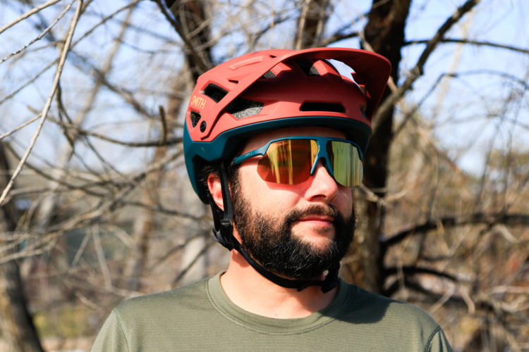 A person wearing a red and teal mountain biking helmet and reflective sunglasses stands outdoors, with a backdrop of bare trees. The individual has a beard and is wearing a green t-shirt.