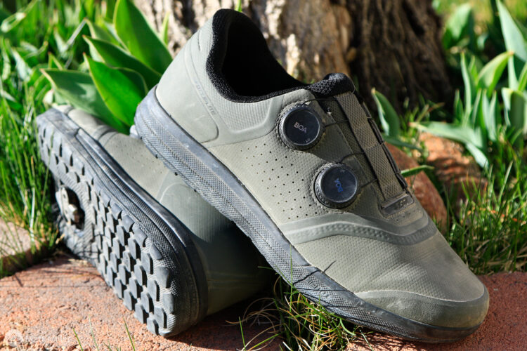 A pair of green athletic shoes featuring a sleek design and two dial adjustments on the side, positioned on grass and rocky ground.