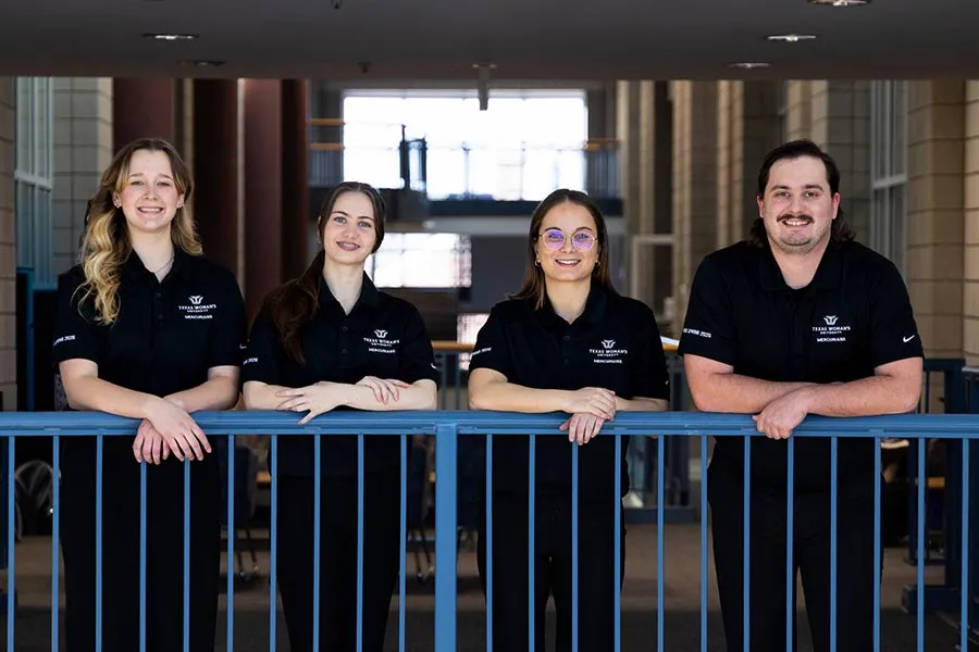 four students in black polos stand behind railing on second floor of Pioneer Hall