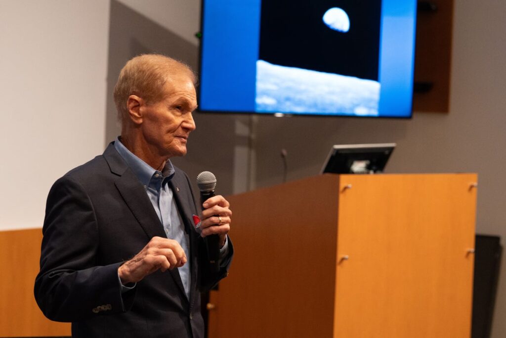 Bill nelson speaking at a podium with the moon displayed on a TV screen behind him