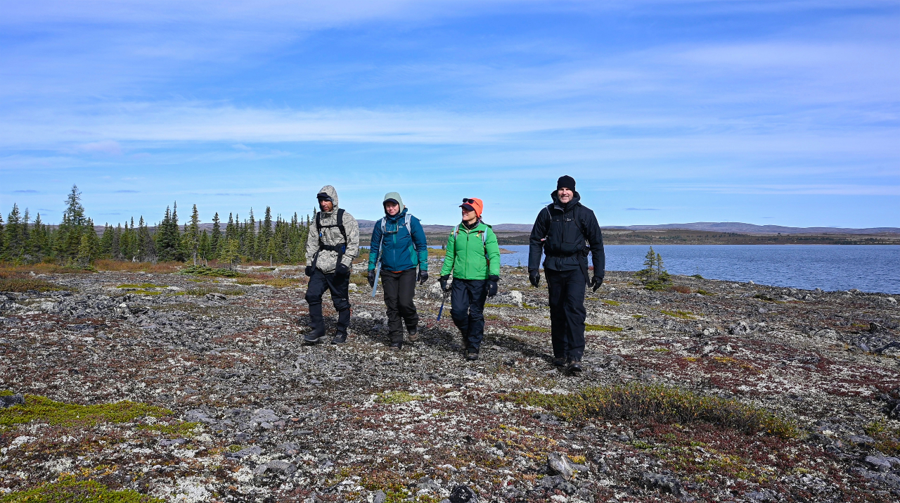 Four individuals wearing large coats walk on a mossy surface with a large lake and forest behind them.
