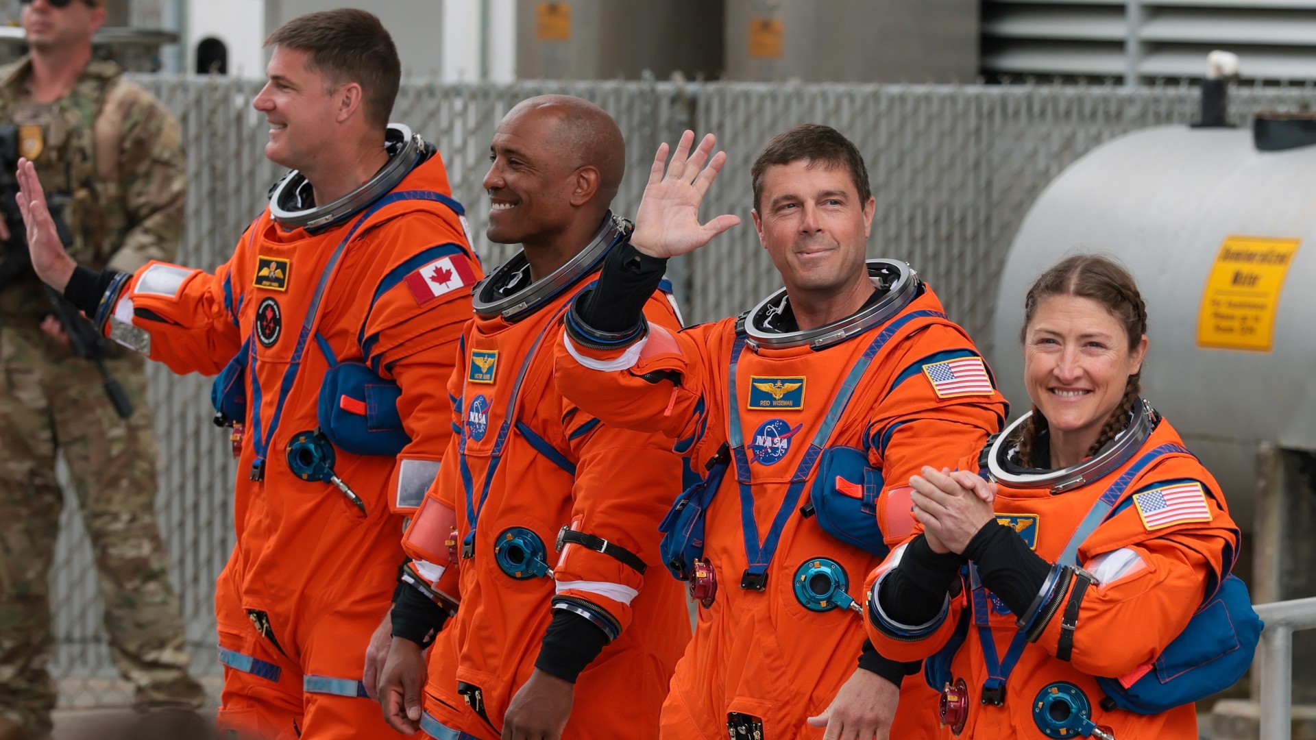 four people in orange flight suits smile and wave to a crowd outside an industrial building