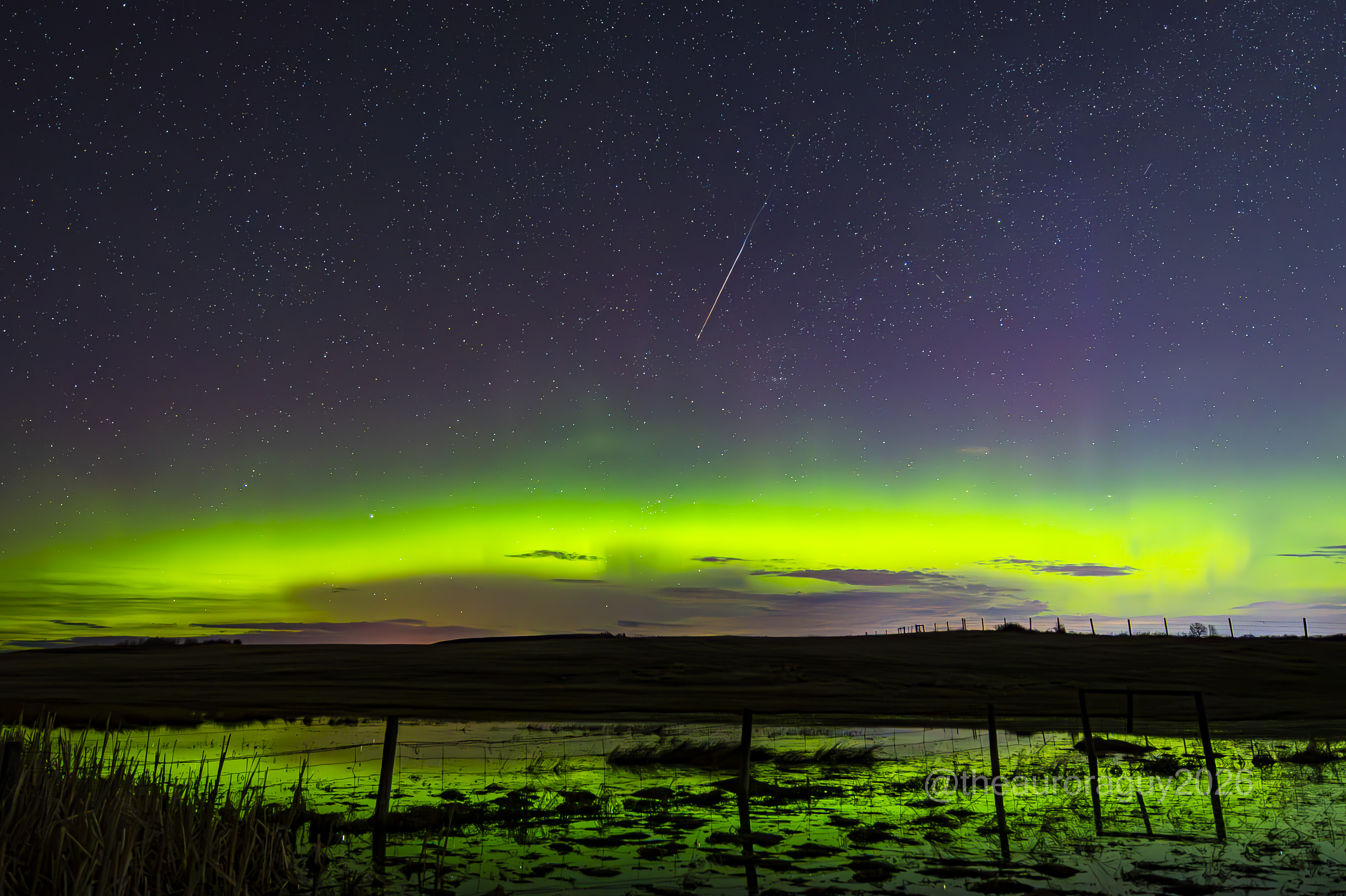 Bright green auroras glowing in a starry sky are reflected in a foreground pond as a meteor streaks earthward at the middle top of the photo.
