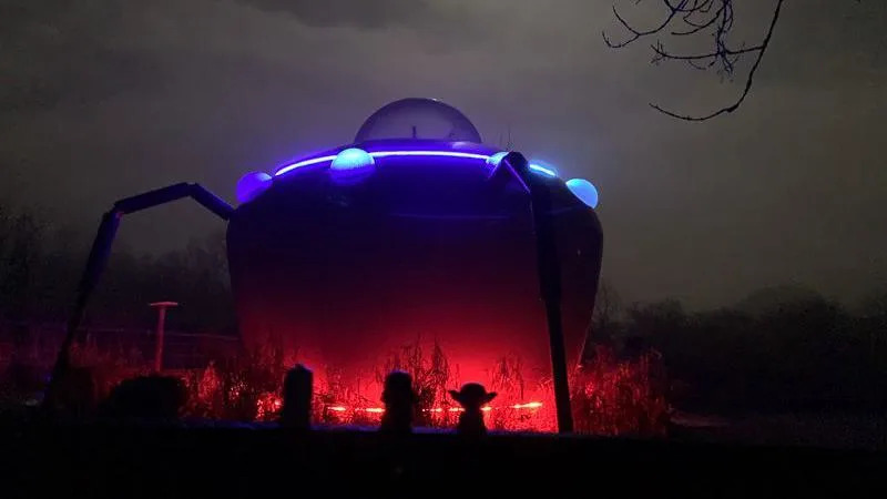 A UFO building lit up in red and blue. Sat in front of a dark moonlit background. Black silhouette of tree branches hang from the top-right corner. 